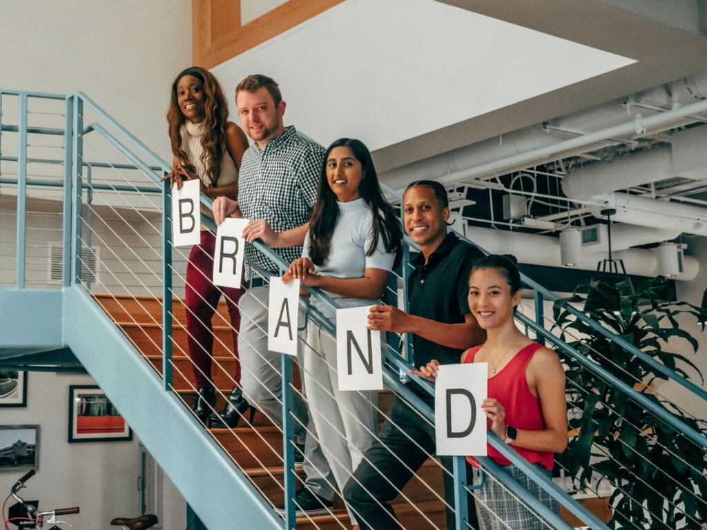 pexels photo 7688457 7688457 A multicultural group of colleagues smiling while holding letter signs spelling 'BRAND' on an office staircase.